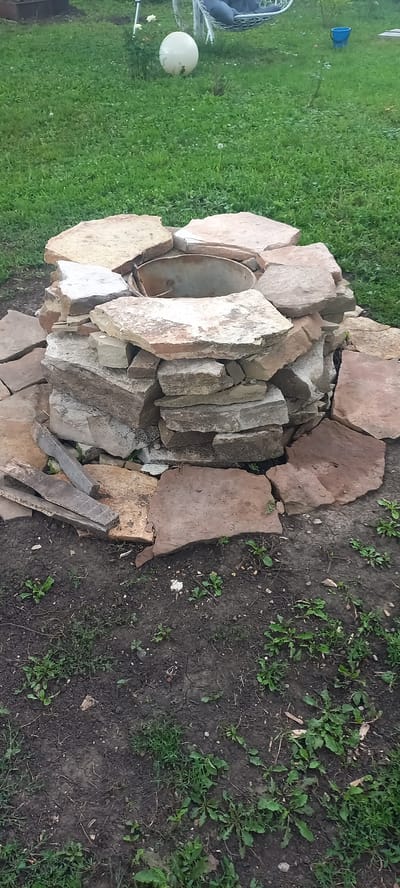 A fire pit constructed from stacked, irregularly shaped stones is visible in a grassy outdoor area, likely a backyard. The stones vary in color, ranging from light tan to reddish-brown, and are arranged to form a circular structure with a metal basin at its center. The ground around the fire pit is composed of dark soil with sparse green vegetation. In the background, out of focus, a white hammock hangs from a stand, a large white spherical object, and a blue bucket are present. The scene suggests a domestic outdoor setting.