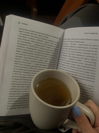 The image depicts a close-up, high-angle view of an open book and a mug of tea resting on a person's lap, covered by dark fabric. The book, open to page 326, displays the title "SAPIENS" and Cyrillic text discussing topics such as human longevity, diseases like Alzheimer's and cancer, and 19th-century medical procedures including amputation and anesthesia. A light-colored, speckled ceramic mug containing a translucent amber liquid, presumably tea, with a visible tea bag string, is held by a right hand. The fingernails of the hand are manicured and painted a solid light blue. The background is out of focus and dark, indicating an indoor environment. The specific location within the city of Lazeshchyna, Ukraine, cannot be determined from the visual information presented in the image.