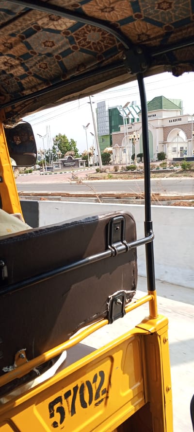 View from inside a yellow auto rickshaw, also known as a Keke Napep, en route to Manhattan Gardens in Uke, Nigeria. The rickshaw's interior features a patterned ceiling, brown seat, and the number "5702" stenciled on the yellow exterior. Visible through the open-air frame is the entrance to Manhattan Gardens, marked by a sign and a gateway structure, as well as a modern building with green accents. Streetlights line the road, and vegetation is seen in the background.