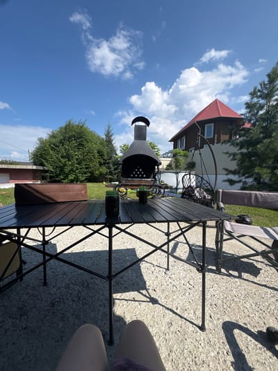 The image depicts a backyard scene on a sunny day in Dzerzhinsk, Russia. In the foreground, a black folding table is set up on a concrete patio, with two folding chairs placed beside it. A brown fabric storage container rests on the table. Behind the table, a large black barbecue grill with a chimney stands on a grassy area. Further in the background, a two-story wooden house with a red roof is visible, along with a swing chair, a white fence, and lush green trees. The sky is bright blue with scattered white clouds. The scene suggests an outdoor recreational space, possibly for a gathering or cookout.