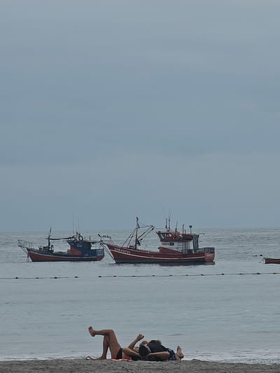 This image depicts a serene coastal scene in Iquique, Chile, under a clear, light blue sky on a bright day. The calm, light blue-grey water stretches from the horizon to a narrow strip of sandy or gravelly shore in the foreground on the right side.

Anchored in the tranquil waters are at least two distinct fishing boats. In the mid-ground, a prominent red and white fishing boat displays the name "DONAVIVI" and the number "1521" on its bow. The registration number "CB3259" is clearly visible on its cabin. Slightly further back, a second fishing boat, with a blue hull and red upper structure, also features the number "1521" and the registration "CB3259" on its cabin, and the name "YOUNIQUE" on its bow. A string of small buoys connected by a rope extends across the water, separating the boats from the shore. A smaller, light-colored boat is faintly visible further out in the distance.

On the sandy shore in the bottom right corner, two individuals are relaxing. One person, largely obscured, wears dark clothing and appears to be sitting. Beside them, a woman in a dark bikini bottom and a lighter-colored top is seen lounging comfortably, with her bare legs and arms visible. Both individuals appear to be enjoying the peaceful atmosphere, looking out towards the sea and the anchored vessels. The scene conveys a sense of leisure and calm in a working coastal environment.