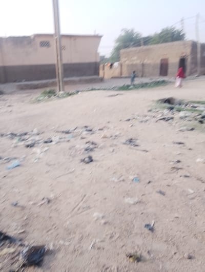 The image depicts an unpaved, sandy street or open area in what appears to be a residential neighborhood in Chiranchi, Nigeria. The ground in the foreground and middle ground is heavily littered with various debris, including plastic bags, papers, and unidentified waste, suggesting poor waste management.
In the blurred background, several single-story, light brown buildings are visible. To the left, one building features a darker brown lower section and a small barred window. To the right, another building shows two dark rectangular openings, possibly doors, and some lighter items that might be laundry hanging outside. Sparse green vegetation is present behind these structures.
A tall, dark utility pole stands prominently on the left side of the frame, extending vertically. Two human figures are in the mid-ground: one, possibly a child, stands to the right of the utility pole, and another, an adult or older youth in red/maroon clothing, walks away to the far right. The lighting suggests early morning or late afternoon, under a diffused, bright sky, with no apparent adverse weather conditions. The overall scene is a typical, albeit cluttered, local street environment. No visible text is present in the image.
