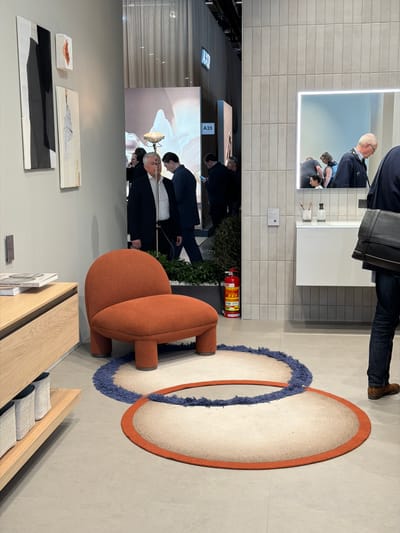 The image captures an indoor exhibition setting at the Rho, Italy location. A red-orange colored, rounded chair sits atop two circular rugs, one with an orange border and the other with a blue, fringed edge. Behind the chair is dense foliage and a red fire extinguisher. Adjacent to the chair is a light wood cabinet with grey cushions. On the wall, artwork is displayed including a black and grey abstract piece, a textured white piece, and a small, red-toned rectangular art object. Above the artwork hangs a large screen showing multiple people in a medical setting. Further up the wall is a rectangular lighted mirror with people reflected inside. A person in a navy jacket and brown shoes is partially seen in the upper right corner, their feet on the tile flooring. A section of tiled wall is visible behind the mirror, and a small label with "A35" is seen on the wall near the screen.