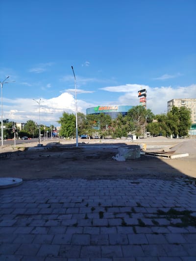 A sunny day is captured in this wide shot of a street scene in Temirtau, Kazakhstan. The sky is a vibrant blue with scattered white clouds. In the foreground, a paved walkway leads to a dirt area with construction materials, including large concrete blocks and metal sheeting. The ground is uneven, suggesting ongoing work.

Further down the street, a modern, multi-story building with a large blue facade is visible. This building features prominent signage reading "KORZINA" in large green letters and "DNS" on a tall tower to its right. The "DNS" tower also displays other advertisements. Surrounding the building is a line of lush green trees.

On the street, several cars are driving, and a few people can be seen walking. Streetlights and traffic signals are also present, indicating this is a busy urban area. To the right of the main building, a taller, older residential building with multiple windows stands out against the sky.

The overall impression is one of a typical day in a developing urban environment, with signs of construction alongside everyday city life. The bright sunlight suggests it is daytime, likely late morning or afternoon. The weather appears clear and pleasant.