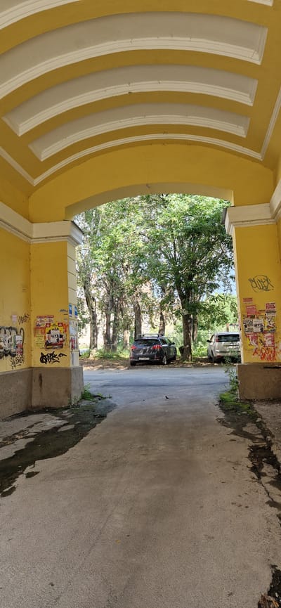 The image shows an archway with yellow walls and a white arched ceiling. Graffiti is visible on the left wall, including stylized lettering and what appears to be a tag. To the right of the archway, more graffiti and posters are affixed to the yellow wall. The ground inside and outside the archway is paved.

Through the archway, a paved road leads to a park-like area with several mature trees. Two cars are parked on the right side of the road: a dark-colored hatchback with a roof rack and a lighter-colored hatchback. The dark car has a Russian license plate. A small bird is perched on the road in the foreground. The overall impression is of an urban environment, possibly an entrance to a courtyard or park in Miass, Russia, characterized by aging architecture, street art, and parked vehicles.