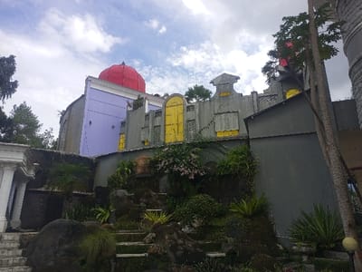 An upward-angled view captures a distinctive multi-level building complex in Gadog, Indonesia, under a partially cloudy sky. The central structure features a prominent red dome atop a lilac-colored wall section, adjoined by a gray facade with white trim. To the right, a separate section of the building displays gray walls accented with yellow, geometric-patterned windows or decorative panels, and castellated rooftop elements. The foreground presents a rugged, landscaped incline composed of large, dark rocks, integrated steps, and dense green foliage, including ferns, shrubs, and climbing vines. On the far left, a white classical architectural element with visible columns and a cornice is partially depicted. A tall, slender tree trunk is visible along the right edge, while the tops of other trees are seen on the left and above the buildings. No individuals or specific actions are discernible in the scene.