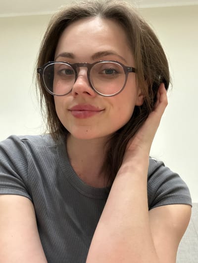 A close-up, eye-level indoor shot of an adult female subject in Scafati, Italy, facing the camera. The subject has fair skin, brown hair, and wears round, dark-framed eyeglasses. She exhibits a slight smile with closed lips. Her right hand is positioned with fingers touching her hair near the right side of her neck. She is wearing a ribbed, short-sleeved gray shirt. The background consists of a plain, light-colored wall, with a gray, possibly wood-grained surface visible in the lower right corner.