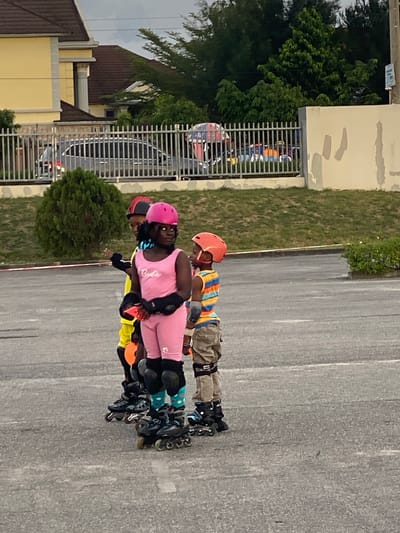Three individuals are inline skating on a paved surface. The first skater wears a pink helmet and a pink outfit with "Barbie" printed on the shirt, as well as elbow and knee pads. The second skater wears a yellow top and an orange helmet. The third skater wears a striped shirt, brown pants, and an orange helmet. They all wear inline skates. The background includes a building, a fence, and some greenery. The image was taken in Sangotedo, Nigeria.