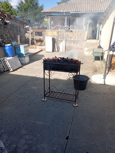 In Buțeni, Moldova, a rustic outdoor scene unfolds with a barbecue grill smoking in the foreground. The grill, a dark metal structure with a decorative base, is filled with what appear to be pieces of corn on the cob, some charred. Smoke rises from the grill, indicating active cooking. To the left of the grill, a large blue plastic barrel and two stacked blue and white beehives are visible, alongside a white radiator. In the background, a weathered wooden structure with a corrugated roof stands, partially obscured by trees. A doorway with steps leads into the building, where more smoke is faintly visible. Various other objects, including wooden pallets, a wheelbarrow, and a black bucket, are scattered around the yard, suggesting a rural or semi-rural setting. The ground is paved with concrete. The sky above is a clear blue.