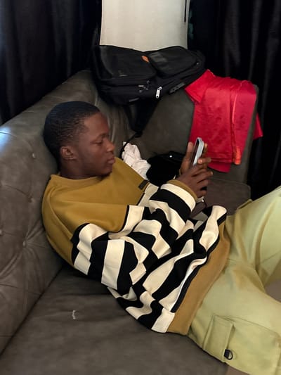 A man is lying on a grey leather couch, looking at a phone. He is wearing khaki pants, a mustard yellow sweatshirt, and a black and white striped sweater. Next to him is a red duffel bag and a black backpack. The environment appears to be indoors. There is no indication of Ikot Ekpene, Nigeria.