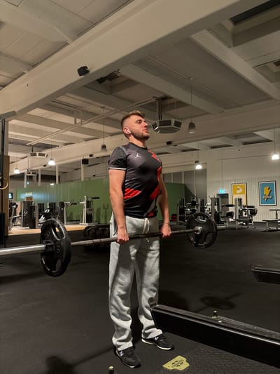 A man with short brown hair and a beard, wearing a black t-shirt with red and white accents and grey sweatpants, is performing a deadlift in a gym. He is holding a barbell with two weight plates on each side, which appears to be a standard Olympic barbell. His stance is centered, and he is looking upwards. The gym environment features a dark rubber floor, weightlifting equipment including machines and a large tire in the background, and a green wall with vertical slats. Overhead, exposed ceiling beams and lighting fixtures are visible. The gym is located in Mönchengladbach, Germany.