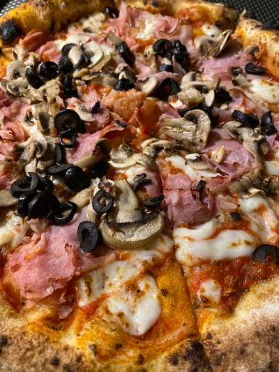 A close-up, overhead, detailed shot shows a freshly baked pizza with a golden-brown crust. The pizza is topped with melted white cheese, thinly sliced ham, black olives, and mushrooms. The toppings are spread evenly across the pizza. The crust has some charred bubbles, indicating it was cooked at a high temperature. The pizza appears to be cut into several slices. The image does not contain any information or clues that would allow for the determination of its location within the city of Banjar, Indonesia.