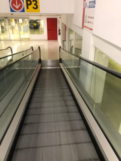 The image displays an empty, indoor moving walkway, also known as a travelator, stretching into the distance. The travelator features a grey, segmented tread and is bordered by clear glass railings with dark handrails. The scene is brightly lit, indicating an indoor commercial or public space, potentially a shopping mall, airport, or large building with access to parking.
On the left side of the frame, a prominent yellow square sign with a black border displays "P3" in large black lettering, likely signifying "Parking Level 3." Below it, a circular red and white sign with a right-pointing arrow guides direction.
In the background, a white wall is visible to the left of the travelator. A bright red cabinet, consistent with a fire extinguisher enclosure, is mounted on this wall. Further back, a section with shelves and what appears to be equipment or display items, including a black object, can be seen. Above the red cabinet, on the white wall, there are some indistinct signs with red and blue elements, but the text is not clearly decipherable.
The perspective looks along the length of the travelator, which appears clean and well-maintained. No people are present, lending a quiet and orderly atmosphere to the modern facility located in Marousi, Greece.
