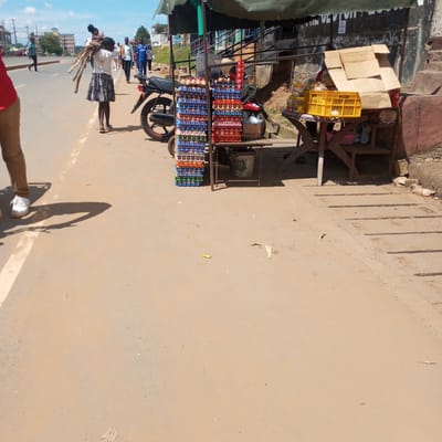 The image shows a street scene in Sheywe Ward, Kenya. On the side of a street, there is a roadside egg stand stacked high with crates of eggs. A motorcycle is parked nearby. A wooden table next to the egg stand holds other items for sale, including a yellow plastic crate and cardboard boxes. A young girl carries a bundle of sticks on her back. Other people are walking in the background. The road surface is rough and appears to be made of packed earth or asphalt.