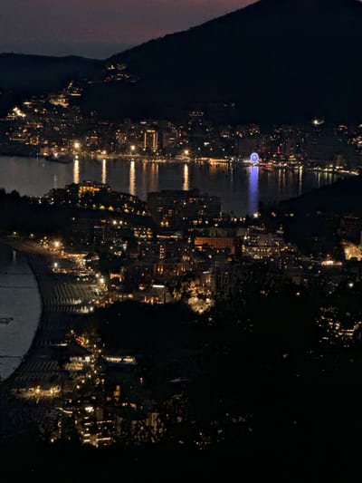 The image is a nighttime aerial view of Podbabac, Montenegro, overlooking a bay lined with a town that is illuminated by numerous lights. A large hill or mountain dominates the background, partially obscuring the sky, which exhibits a subtle gradient of dark purples and blues.
The town features a dense collection of buildings with lit windows, suggesting residential and commercial activity. Along the water's edge, several boats are visible, their lights reflecting on the dark water. A prominent Ferris wheel, illuminated with blue lights, stands out in the middle distance, a focal point of the cityscape. The coastline is also marked by a beach area with rows of what appear to be sun loungers or umbrellas, many of which are also lit. The foreground consists of dark foliage, silhouetted against the illuminated town and water, suggesting the vantage point is from a higher elevation on a hillside. The overall impression is one of a vibrant, bustling town at night, nestled beside a tranquil body of water and dramatic natural landscape. There are no people visible in the image.