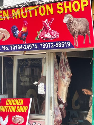 The image shows a storefront in Dharamkot, India, for a "Chicken Mutton Shop." The exterior features a red sign with yellow lettering advertising the shop's name and two phone numbers: 70184-24974 and 78072-58519. The sign also displays images of a rooster, raw meat, and a sheep. Inside the shop, carcasses of meat hang from the ceiling. A person's hand is visible pointing towards the hanging meat. In the background, a reflective window shows a partial view of what appears to be a sign with the words "YOUR TRIP STARTS HERE." The scene suggests a typical local butcher shop in the region.