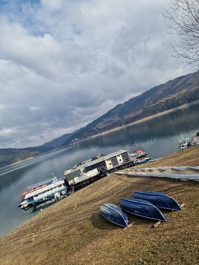 This image captures a wide view of Lake Izvorul Muntelui (Bicaz Lake) in Bicaz, Romania, under a partially cloudy sky. In the foreground, a dry, sloping grassy bank, showing patches of brown earth, occupies the lower right portion of the frame. Three blue, inverted small boats are positioned parallel to each other on this bank. Along the waterline, two larger vessels are moored or docked: a multi-level passenger boat, primarily white and blue with red trim, and behind it, a long, two-story structure resembling a floating building or barge, dark gray and white. Further along the shore to the right, a smaller green and white boat is docked. The calm water of the lake extends towards a range of forested hills and mountains that rise in the background under a mix of overcast and blue sky. A bare deciduous tree branch is visible in the upper right corner of the frame. No individuals are present in the scene.