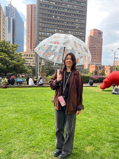 A young woman stands on a green lawn holding a clear umbrella with dog designs. She wears a brown jacket, a black top, dark jeans, and black shoes. A pink phone hangs from her neck. Tall buildings are visible in the background, along with a red sculpture. Other people are seated nearby on a ledge. The sky is cloudy, suggesting a partly overcast day.