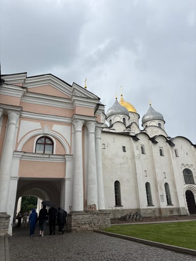 The image depicts the Saint Sophia Cathedral in Veliky Novgorod, Russia. The cathedral, a prominent landmark, features white walls, grey domes, and a single golden dome, topped with crosses. A light pink structure with classical columns is attached to the left side of the cathedral, forming an archway. Several people are walking under the archway, with some holding umbrellas, suggesting it is raining. The ground is paved with cobblestones, and a strip of green grass is visible in front of the cathedral. The sky is overcast and cloudy.