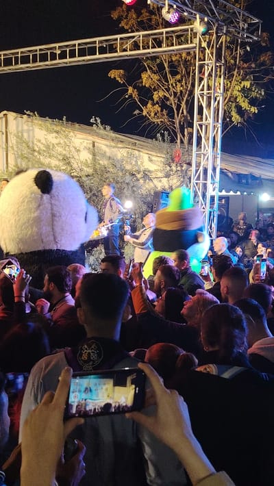 A large crowd gathers at night, many holding phones to record a performance. A man on a raised platform plays an instrument while another films him. To their left stands a person in a giant panda costume, and to their right, a person in a green and brown costume. The stage has metal scaffolding with colored lights. Trees and buildings form the backdrop to the stage, illuminated by the stage lighting.