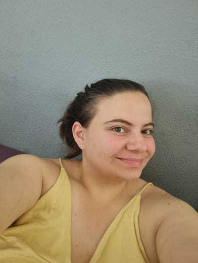 A selfie photograph shows a woman with fair skin and brown hair pulled back away from her face. She is facing toward the viewer with a slight smile. She is wearing a yellow tank top with thin straps. A wall of textured, light-blue material is behind her. The image doesn't provide enough information to place the subject in Saarbrücken, Germany.