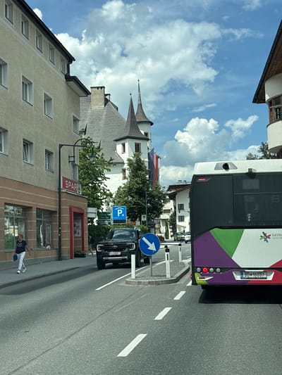 This image captures a street scene in Zell am See, Austria, on a sunny day with a partly cloudy sky. In the foreground, a large bus is partially visible on the right, featuring a colorful design with purple, green, and white sections, and the text "Salzburg verbunden". The bus has a license plate reading "BD 16037". A black SUV with the license plate "ZE HOHKR 1" is in the middle lane, facing forward. To the left of the SUV, a blue circular sign with a white arrow pointing downwards is visible, indicating a directional instruction.

On the left side of the street, a yellow building with multiple windows is present. A "SPAR" supermarket is located within this building, with its red and white signage clearly displayed. A street lamp with a green curved arm and two lamps extends over the sidewalk. A woman in a dark top and white pants is walking on the sidewalk, carrying a blue bag. Further down the street, several other buildings and trees are visible, including structures with distinct spires that are characteristic of Zell am See. The road has clear lane markings, indicating a multi-lane street. The overall environment suggests a typical urban setting with shops, traffic, and pedestrian activity.