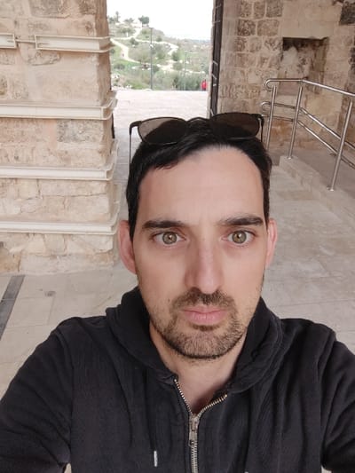 A male individual is centered in a close-up, frontal photograph, looking directly at the camera with a neutral expression. He has dark hair, a short, trimmed beard, and light green-hazel eyes. He is wearing a dark, zippered hooded sweatshirt, with dark sunglasses resting on top of his head.

The subject is positioned within or immediately in front of a stone architectural structure, likely a passage or large doorway, in Rosh HaAyin, Israel. The structure is constructed from light-colored, rectangular cut stone blocks. To the left, a stone pillar exhibits several horizontal, white, planar protrusions. To the right, a stone wall features a recessed niche and a flight of light-colored steps accompanied by a silver-colored metal handrail.

Beyond this structure, an outdoor park environment is visible. This includes paved pathways traversing a gently sloping, vegetated landscape with scattered trees and lampposts under an overcast sky. A distant hill with faint structures is present on the horizon. A small, partially obscured figure in red clothing is discernible in the mid-ground, near a pathway. The ground in the immediate foreground where the subject stands is a light-colored paved surface.