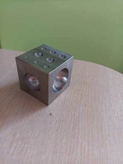 A close-up, slightly elevated shot shows a metal cube with circular holes on its top and sides, resting on a light-colored, wood-grained surface. The cube is positioned to the left of the frame, with its top surface facing the camera and slightly angled. Three circular indentations are visible on the top surface, varying in size. Two large, circular openings are visible on the front and right sides of the cube, revealing reflections within their hollow interiors. The background consists of a solid, muted green wall. The lighting appears to be natural or ambient, casting soft shadows. The image is crisp and in focus, emphasizing the texture of both the metal cube and the wooden surface. The scene is devoid of any human presence or activity. The location of Vilyuysk, Russia, is not ascertainable from the visual information provided in the image.