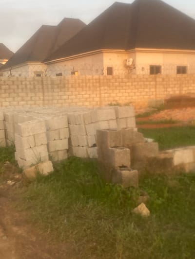 The image displays a residential development within Iddo Tudunwada, Nigeria. In the background, several houses feature dark, pitched roofs and light-colored exterior walls. One house exhibits an externally mounted air conditioning unit and multiple rectangular windows. A decorative wall with security spikes or barbed wire is visible along the base of these structures. A substantial, unplastered concrete block wall forms a boundary across the midground. In the foreground, numerous stacks of light-grey concrete blocks, consistent with construction materials, are arranged on patches of green grass and exposed reddish-brown soil. A dirt path is present in the lower-left corner. Scattered concrete debris lies on the ground. The lighting indicates either early morning or late afternoon. No individuals are present in the scene.