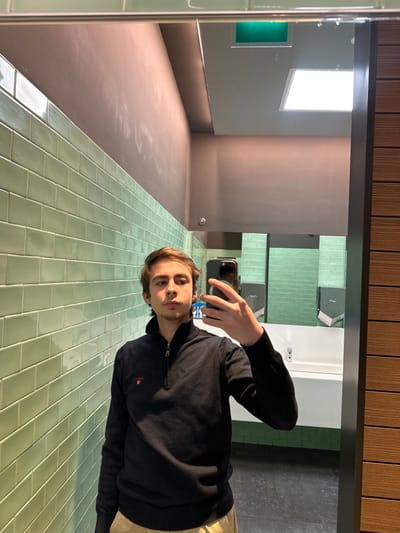 A mirror reflects a young man taking a selfie in a public restroom. He wears a dark blue zip-up sweater and khaki pants, holding his phone in his right hand. The restroom features light green tile on the walls and floor. Reflected in the mirror is a minimalist space with a large window, a green rectangular panel, a sink, and toilet paper dispensers. Above the mirror is a wood-paneled ceiling.