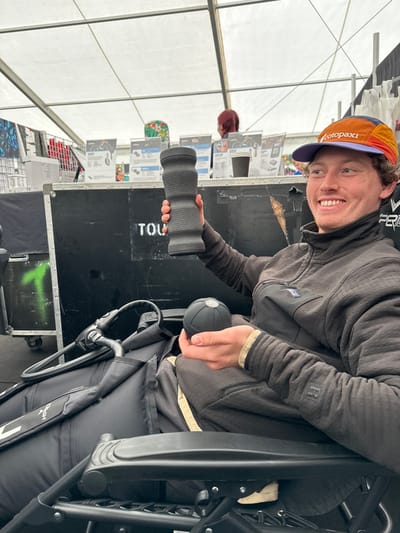 An adult male with light skin and short brown hair is seated within a large white canvas marquee tent. He wears a multi-colored "Cotopaxi" baseball cap, a dark textured "Patagonia" fleece jacket with an "R" logo on the right sleeve, and dark trousers. He is actively smiling towards the camera while holding a black, textured, cylindrical massage roller (similar to a Theragun Wave Roller) aloft in his right hand. In his left hand, he holds a black, textured, spherical massage ball (similar to a Theragun Wave Solo). He is seated in a black adaptive sports chair or racing wheelchair, which features a black frame and "HyPro" text visible on a strap.
In the background, a display booth is set up with black modular walls or crates. Various product information cards or flyers are displayed on stands, alongside a white paper coffee cup. Green spray paint marks and partially stenciled white text, including "TOU" and "FER," are visible on the black display surfaces. A person with red hair is partially visible standing behind the display.
The scene depicts an event village or expo setting, consistent with an endurance sports event such as IRONMAN 70.3 Weymouth, situated in Weymouth, United Kingdom.