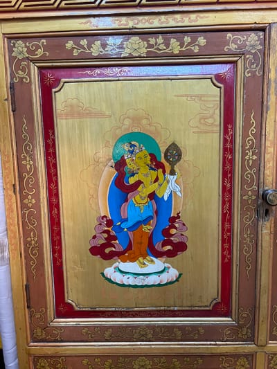 The image shows a detailed decorative panel, likely from a piece of furniture or architecture. The panel features a gold-colored background with an intricate painting of a figure resembling a deity or spiritual being from Tibetan Buddhism or a related tradition. The figure is depicted with yellow skin, ornate jewelry, and flowing garments in shades of blue, red, and white, seated on a lotus platform. The background includes stylized cloud motifs. The panel is framed with multiple layers of decorative borders in gold, red, and brown. The borders include floral and geometric patterns. The overall aesthetic suggests a cultural context associated with Tibetan Buddhism, placing it potentially in the city of Dharamshala, India, known for its Tibetan community and Dalai Lama residence. The image contains no people.