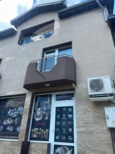 The image shows the facade of a building, likely a commercial establishment, in Beloslav, Bulgaria. The building has a light-colored stucco exterior with dark brown trim and accents. A small balcony with glass railings juts out from the second story. On the ground floor, there are large display windows and a doorway. One window displays images of automotive parts and the word "АВТО" (Avto), indicating an auto-related business. The other window and the doorway showcase various brands of car parts, oils, and additives, with the word "ДОБАВКИ" (Dobavki) also visible, meaning additives. Several logos of car manufacturers like Renault, Audi, BMW, and Nissan are displayed. An outdoor air conditioning unit is mounted on the wall to the right of the entrance. The sky above is partly cloudy. The image is taken from a low angle, looking up at the building.