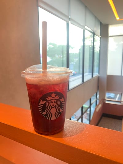 A cold Starbucks beverage in a clear plastic cup with a straw sits on an orange railing. The cup has the Starbucks logo visible. The background shows a brightly lit indoor space with large windows overlooking an outdoor area with trees and what appears to be a city street. The railing is smooth and painted orange. The lighting suggests it is daytime. There are no people in the frame. This scene could be a cafe or a seating area within a building, possibly in a commercial district in Angeles City, Philippines, given the architectural style and general urban environment suggested by the background.