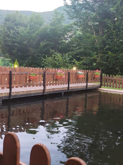 The image depicts a tranquil outdoor scene with a focus on a wooden walkway extending over dark water. The walkway is bordered by a rustic wooden fence, and several planters with pink and white flowers are attached to the fence. Two small lights are visible on the fence posts, suggesting it is evening or dusk. Behind the fence, a dense forest of green trees covers rolling hills, fading into a hazy sky. The water in front of the walkway is still, reflecting the fence, walkway, and the surrounding trees, with subtle ripples distorting the reflections. In the foreground, the tops of two wooden fence posts are visible. The overall atmosphere is peaceful and natural, likely from a park, garden, or resort setting. No people are visible, and no text is present in the image.