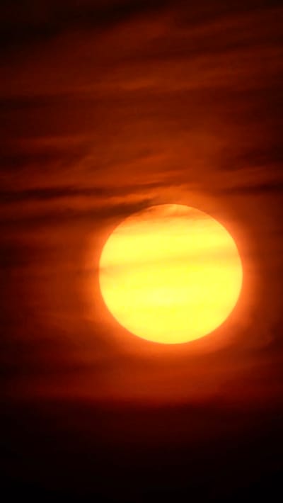 The image shows a close-up of the sun, a large yellow orb, partially obscured by dark red clouds. The clouds are wispy and streaky, creating a dramatic contrast with the bright sun. The scene is dark, with the lower portion of the image fading into black. The overall impression is one of an intense and atmospheric sunset or sunrise. The location is specified as Asan-si, South Korea. There are no people, subjects, or objects other than the sun and clouds depicted in the image.