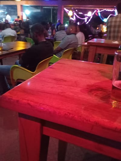 The image shows a close-up of a red wooden table in a dimly lit bar or restaurant. It appears to be situated in Arusha, Tanzania, at the coordinates provided. There are people seated at tables in the background, with some conversing. One individual is seated to the left of the table, facing away from the camera. A glass of beer is partially visible on the table in the foreground. There are a few other objects on the table, including a cell phone. There are also empty beer bottles on the table to the left. The scene is a typical bar or restaurant environment, with a dimly lit interior and a bustling atmosphere. The furniture is mostly made of wood and has a worn appearance.  The walls are decorated with string lights and decorations. The background is out of focus and blurred, and the focus is on the table and the glass of beer. The overall image is reminiscent of a lively night spot.