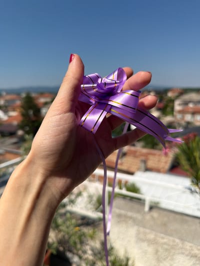 A close-up shot captures a person's hand holding a shimmering purple ribbon with gold stripes. The ribbon is intricately folded into a decorative bow or ornament, with two pointed ends extending upwards like antennae. The hand, with a hint of red nail polish on the thumb, is illuminated by bright sunlight, casting a shadow across the palm.

In the background, a blurred cityscape unfolds under a clear blue sky, suggesting an outdoor setting. The buildings, with reddish-brown roofs and white facades, indicate a warm, sunny day. Lush green foliage and glimpses of what appear to be trees add to the natural element of the scene. The overall impression is one of bright daylight, possibly late morning or early afternoon, given the strong sunlight and shadows. The context of Svilengrad, Bulgaria, places this scene in a European urban environment. No text is visible in the image.