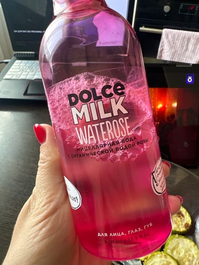 A close-up, eye-level shot shows a person's hand with bright red nail polish holding a clear, pink plastic bottle of "DOLCE MILK WATERROSE" micellar water. The bottle is partially filled with a pink liquid, which has a frothy top layer. Text on the bottle is visible, including "DOLCE MILK WATERROSE" in large black and white letters, and smaller Russian text that translates to "micellar water with organic rose water." At the bottom, it states "FOR FACE, EYES, LIPS 400 ml / 13.5 fl.oz." The hand holding the bottle is positioned in the foreground, with the bottle angled slightly away from the camera. In the background, out of focus, are a laptop and a kitchen appliance, possibly an oven. The scene appears to be indoors, likely a home kitchen environment. The image does not provide any specific visual cues to determine the exact location within the city of Novoaltaysk, Russia.