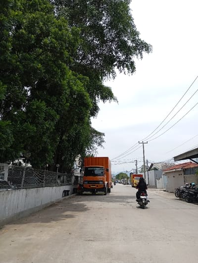 The image shows a street scene in Katapang, Indonesia, taken from the sidewalk looking down the street. The latitude and longitude coordinates are -6.986 and 107.556, respectively. A large, orange, box truck is parked on the right side of the road, facing away from the viewer. Several smaller trucks and cars are visible in the distance. A person is riding a motorcycle down the street, wearing a dark jacket and helmet. The sidewalk is lined by a gray concrete wall, and a large tree with green leaves is in the foreground. The sky is overcast. Power lines run across the scene. The asphalt road surface is clean and dry. There are no other people visible in the scene.