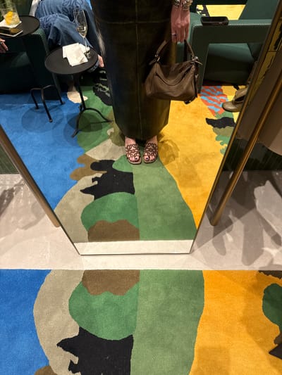 The image presents an overhead view of an indoor lounge or waiting area, likely in Zapopan, Mexico, characterized by a modern and upscale design. The scene incorporates reflections from mirrored surfaces, adding depth and complex visual elements.

**Subjects:** Parts of at least two people are visible. One person's legs and feet, adorned with brown patterned sandals and dark toe nail polish, are prominent, suggesting they are seated on dark green upholstered furniture. Another individual's hands and lower arms are seen holding a distinctive brown leather handbag, possibly a Loewe Puzzle bag, with silver hardware. This person wears multiple silver bracelets and a ring with a purple stone. A third person's legs, clad in blue jeans, are partially visible in the bottom left corner, also seated.

**Objects:** Key objects include a large, abstract rug featuring organic shapes in a vibrant color palette of yellow, various greens, blues, brown, and black. Dark green upholstered furniture serves as seating. Two small, black metal side tables are present; one holds an empty champagne flute and white napkins, while the other holds a glass with ice and a lemon slice, alongside a mobile phone. A long, shiny golden or brass handrail extends across the frame. The surrounding floor is made of light gray or beige tiles, with mirrored sections creating reflections. A black-cased smartphone is also visible resting on the arm of the green furniture.

**Activity:** The individuals appear to be relaxing in a social or leisure setting, possibly enjoying drinks. The presence of the handbag suggests they might be waiting, arriving, or departing.

**Notable Details:** The lighting is characteristic of an indoor environment, possibly evening or late afternoon, with artificial light casting subtle shadows. The overall atmosphere conveys a sense of relaxation and sophistication. No visible text is present in the image.