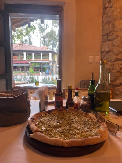 The image depicts a table setting inside a restaurant, likely in Domingo Arenas, Mexico, based on architectural styles visible through the window.
On the white tablecloth, there is a pizza on a dark-colored wooden serving plate, covered with green herbs or spices. Next to the pizza, there's a pair of sunglasses. There are also several bottles of olive oil and various hot sauces arranged in a row. To the lower left is a tan-colored breadbasket with bread inside.
Through the window, a view of buildings and trees is visible, including architectural details suggestive of a Mexican town. The scene is indoors, with stone walls visible behind the table setting.