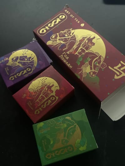 Four boxes of tea are arranged on a dark, flat surface. The boxes are rectangular and feature elaborate, gold-colored designs of horses and stylized clouds. The main box, which appears to be the largest and is partially open, is dark red and has the text "ART OF TEA" printed on it. Two smaller boxes, one purple and one red, are positioned near the main box. A fourth, green box is located in the lower right corner. The arrangement suggests they might be part of a collection or display. The image does not provide any information that would allow for the determination of its location in the city of Kommunarka, Russia.