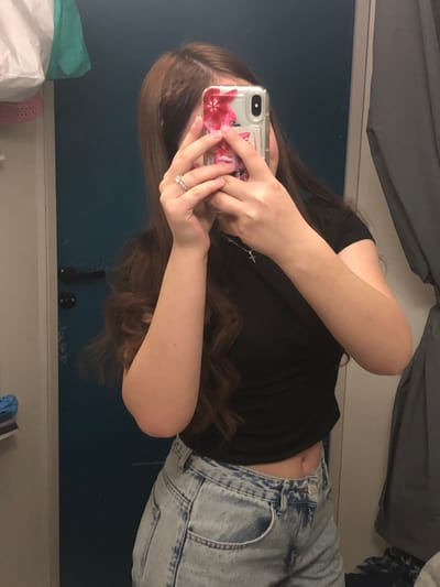 A single individual, appearing feminine, is captured from the mid-torso upward in a mirror reflection within an interior space, consistent with a commercial fitting room, located in the city of Saverne, France. The specific establishment within Saverne is not identifiable from the visual information.

The individual, with long, wavy brown hair, is wearing a black short-sleeved crop top and light-wash denim shorts or jeans, revealing the midriff. A silver-toned necklace with a small cross pendant is visible on the neck. A ring with a clear, faceted stone is worn on the left ring finger, alongside a thinner band. The individual is holding a smartphone with both hands, obscuring the face, in the act of taking a mirror selfie. The smartphone is enclosed in a clear case featuring a pink floral design, and its rear camera module is visible.

The immediate background to the left shows a dark teal-colored door with a black handle. Above the door, light-colored fabric items are hung. To the right, a grey fabric curtain is partially visible, consistent with a changing area. The lighting appears to be artificial indoor illumination. No other persons or interactions are present.