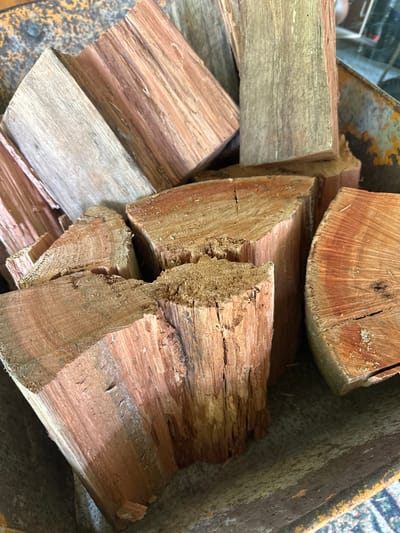 A close-up, overhead shot shows a pile of chopped firewood with a reddish-brown hue, resting inside a weathered, orange and grey metal container, possibly a wheelbarrow, in Thora, Australia. The wood pieces vary in size and shape, with some exhibiting rough, splintered ends and others showing more finished, circular cross-sections. The grain of the wood is clearly visible, displaying variations in color and texture. The container's surface appears worn and textured, suggesting age and use. The lighting is natural, casting soft shadows that highlight the contours of the wood and the container.