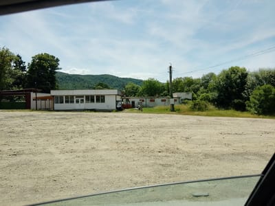 The image shows a tan-colored dirt parking lot in front of a long, white, single-story building with a row of large windows and a covered entrance. Behind the building is a dense line of green trees and hills, under a sky with scattered white clouds. A utility pole with wires stands to the right of the building. The scene appears to be in a rural or semi-rural area, possibly the outskirts of Mirovo, Bulgaria, given the architecture and vegetation.