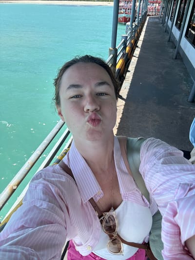 A woman with dark hair tied back, wearing a pink and white striped shirt and a white top, leans forward to take a selfie on a pier in Nathon, Thailand. Her lips are puckered, and she is looking at the camera. Sunglasses hang from her shirt. She is standing next to a railing overlooking turquoise water, with a walkway and boats visible in the background.