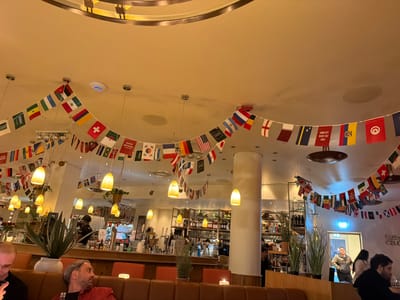 The image depicts the interior of a cafe or restaurant in Kassel, Germany. A multitude of national flags are strung across the ceiling, suggesting a celebratory or international theme. In the foreground, several patrons are seated on a booth. To the left, a man with a shaved head is looking down at his phone. In the center-right, a man with gray hair and a beard is looking to the right, wearing a red jacket. The background reveals a bar area with shelves stocked with bottles and glasses, and a bartender is visible. Warm lighting emanates from pendant lamps throughout the space. A large plant in a pot is positioned near the bar.