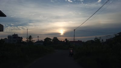 This is a photograph taken at dusk in Uteunreutoh, Indonesia. The sky is filled with a mixture of clouds and the setting sun, which is visible as a bright orb near the horizon, casting a warm glow. There is a subtle sundog visible on the left and right of the sun. A dark road leads into the distance, bordered by dense green foliage on both sides. In the middle ground, several buildings and trees are silhouetted against the sky. A person on a motorcycle is riding on the road, with their headlights illuminated. Electrical wires crisscross the sky, supported by poles. The overall atmosphere is serene and peaceful, indicative of the end of the day.