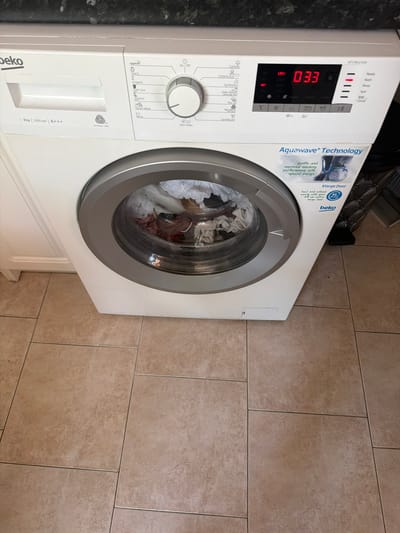 In Novi Iskar, Bulgaria, a white Beko washing machine is positioned on a tiled floor. The washing machine has a load of laundry inside its drum. The control panel is visible, displaying a digital timer showing "033" and various program selection options. The machine features an "Aquawave Technology" sticker and an "XL Door" indicator. The surrounding environment appears to be an indoor domestic setting, possibly a kitchen or utility room, with cabinets visible to the left and a textured dark surface above. The overall scene is static and well-lit.